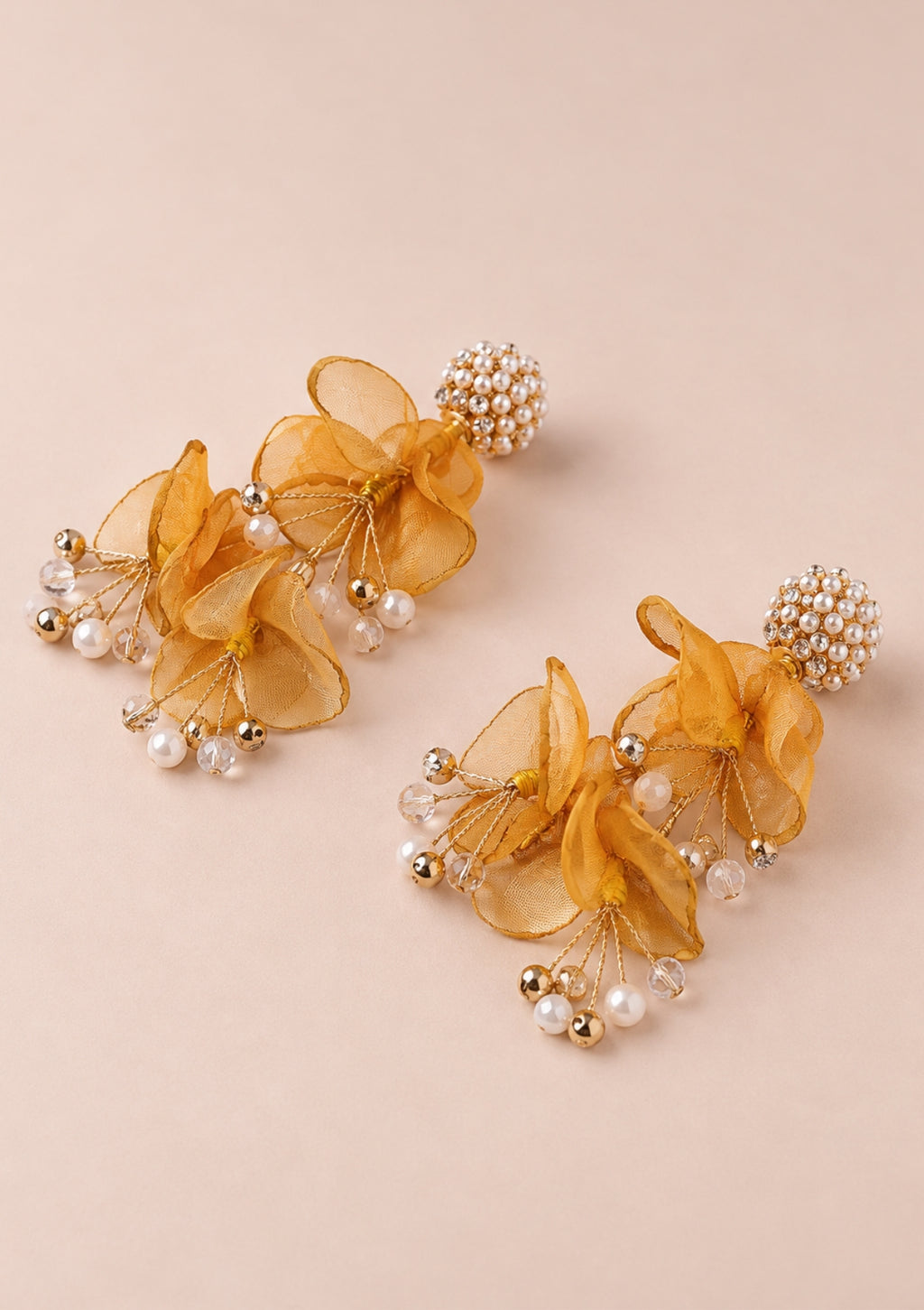 A pair of statement dangle earrings featuring layered sheer golden-yellow organza flowers, small white faux pearls, and gold-toned metallic beads, displayed on a solid pale pink background.