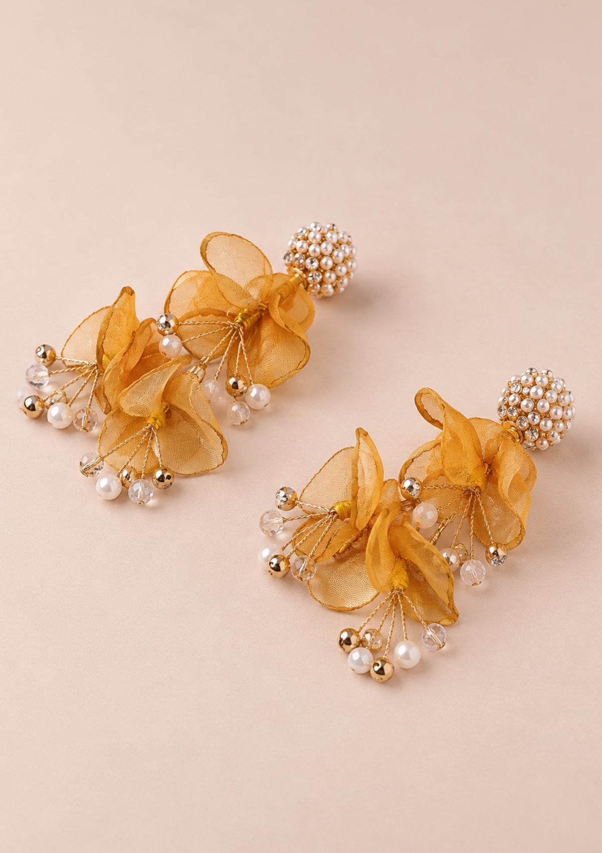 A pair of statement dangle earrings featuring layered sheer golden-yellow organza flowers, small white faux pearls, and gold-toned metallic beads, displayed on a solid pale pink background.