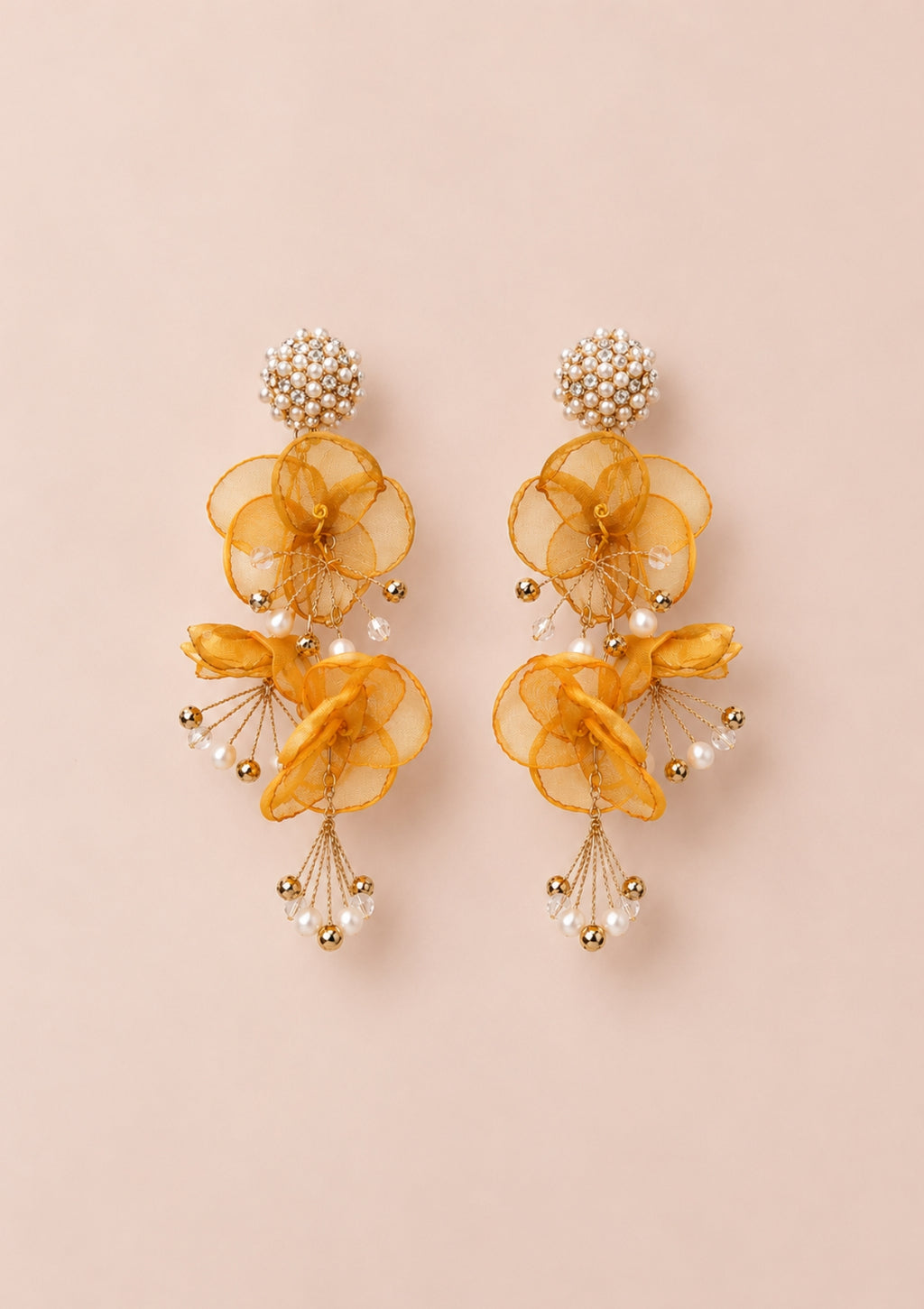 A pair of gold and yellow dangling earrings with layered sheer yellow fabric flower shapes, small pearl-like beads, and gold-tone metal details, displayed on a solid pale pink background.