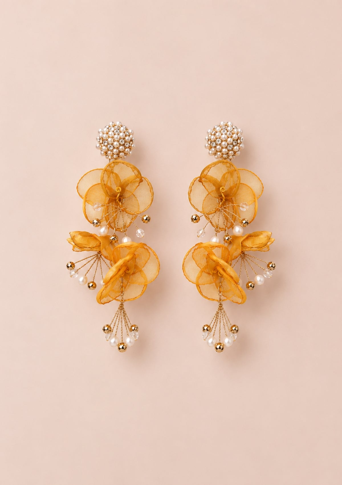 A pair of gold and yellow dangling earrings with layered sheer yellow fabric flower shapes, small pearl-like beads, and gold-tone metal details, displayed on a solid pale pink background.