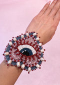 Hand wearing a beaded bracelet with red, blue, and white colors on a pink background