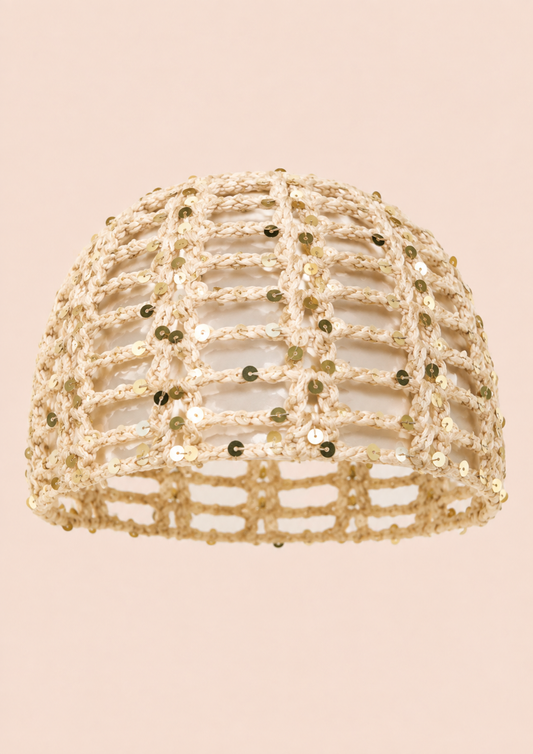 A beige crochet openwork hat embellished with small gold sequins, shown against a solid pale pink background.