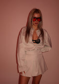 Load image into Gallery viewer, Woman wearing a beige outfit with red sunglasses against a pink background
