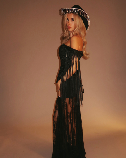 Woman wearing a black off-shoulder coerset with sheer panels and a decorative hat against a beige background