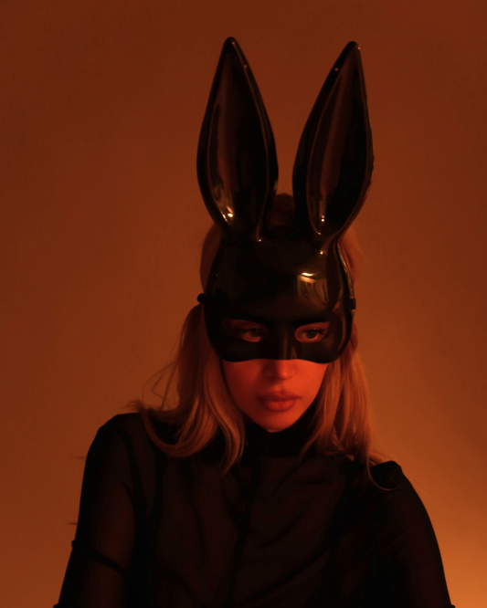 Person wearing a black bunny mask with pointed ears against an orange background