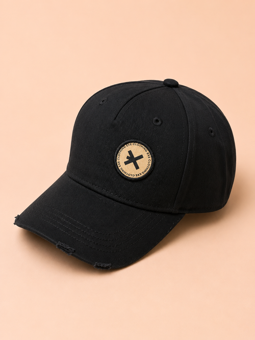 A black distressed baseball cap with a circular beige patch featuring a black 'X' logo on the front, set against a solid peach background.