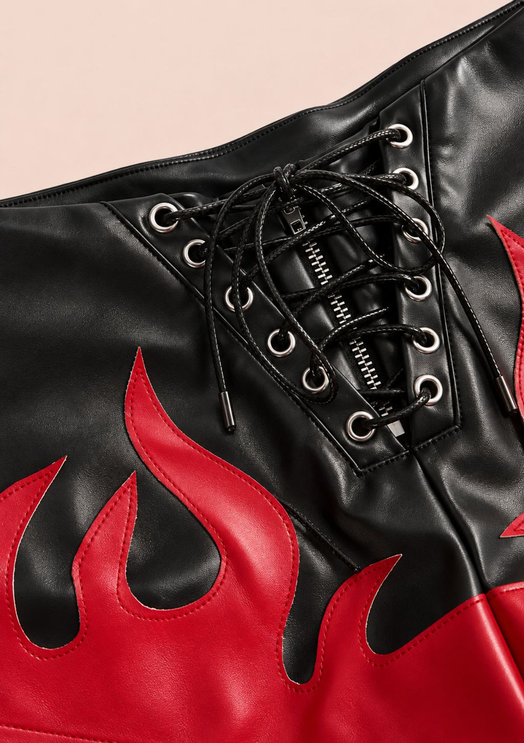 Close-up of a black faux-leather lace-up shorts with red flame applique and silver metal eyelets, shown against a pale pink background.