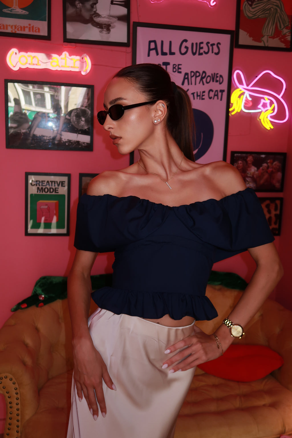 Woman in a black off-shoulder top and white pants standing in a room with pink walls, neon signs, and framed pictures.