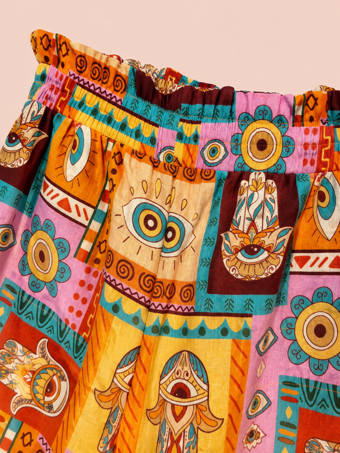 A close-up of a colorful patchwork shorts featuring bohemian Hamsa hand and eye motifs in orange, pink, yellow, teal, and brown on a solid pale peach background.
