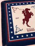 Load image into Gallery viewer, A close-up of a woven throw scaf with fringe, featuring a western rodeo design. The blanket has a cream center with a dark red rearing cowboy silhouette, blue and red bull skulls, and text reading 'EST' and 'THIS AIN'T MY FIRST RODEO'.
