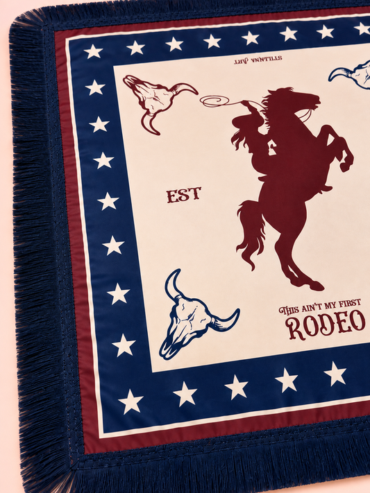 A close-up of a woven throw scaf with fringe, featuring a western rodeo design. The blanket has a cream center with a dark red rearing cowboy silhouette, blue and red bull skulls, and text reading 'EST' and 'THIS AIN'T MY FIRST RODEO'.