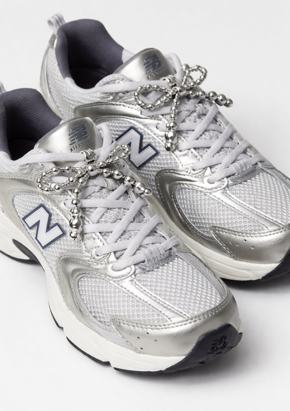 Silver New Balance sneakers with decorative bow-shaped shoe charm on a white background