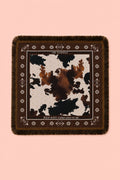 A square fringed scarf with a brown and black cow print center and a dark brown border with geometric patterns, displayed flat against a solid pink background.