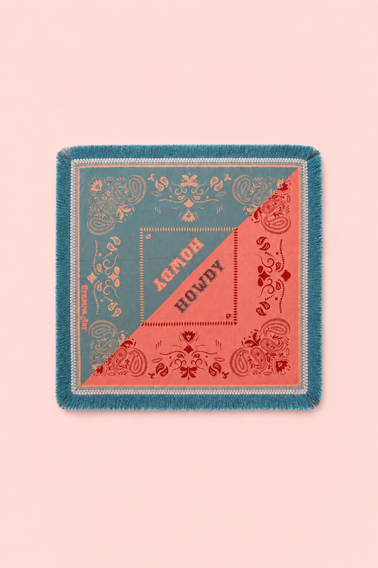 A square fringed scarf laid flat on a solid pale pink background. The scarf features a split design with teal blue on one side and coral pink on the other, decorated with ornate floral patterns and the words 'ALMOST HOWDY' printed diagonally across the center.