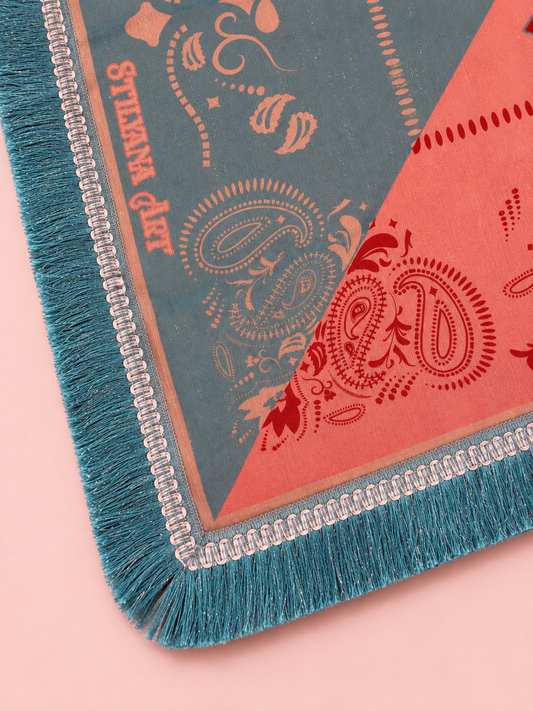 A close-up of a patterned textile or rug corner with teal fringe, a muted blue-gray field, coral pink sections, and ornate paisley and floral motifs. The words 'STAYAWAY' are printed vertically on the blue-gray section.