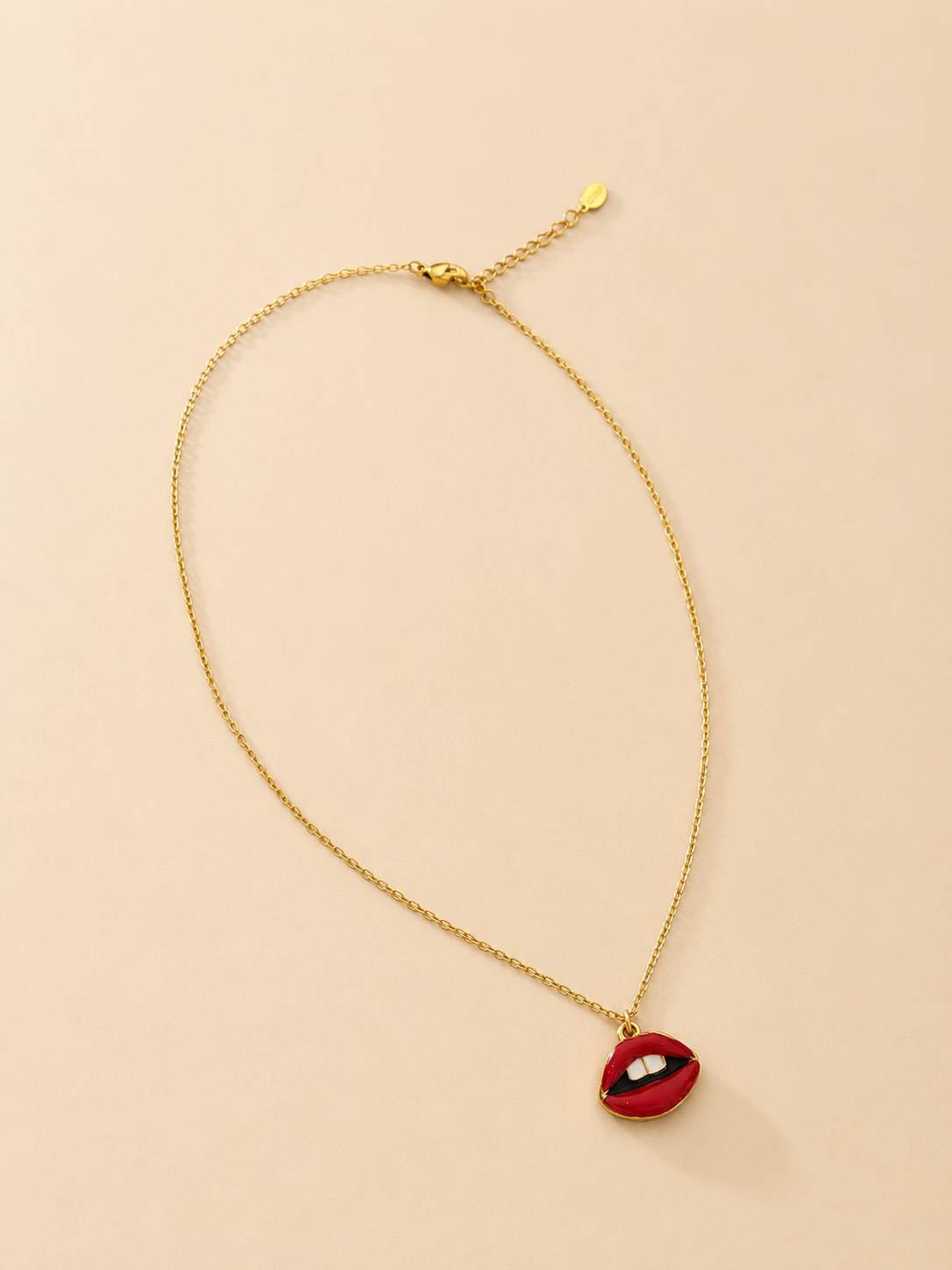 A gold chain necklace with a small red lips pendant featuring white teeth, displayed on a light beige background.