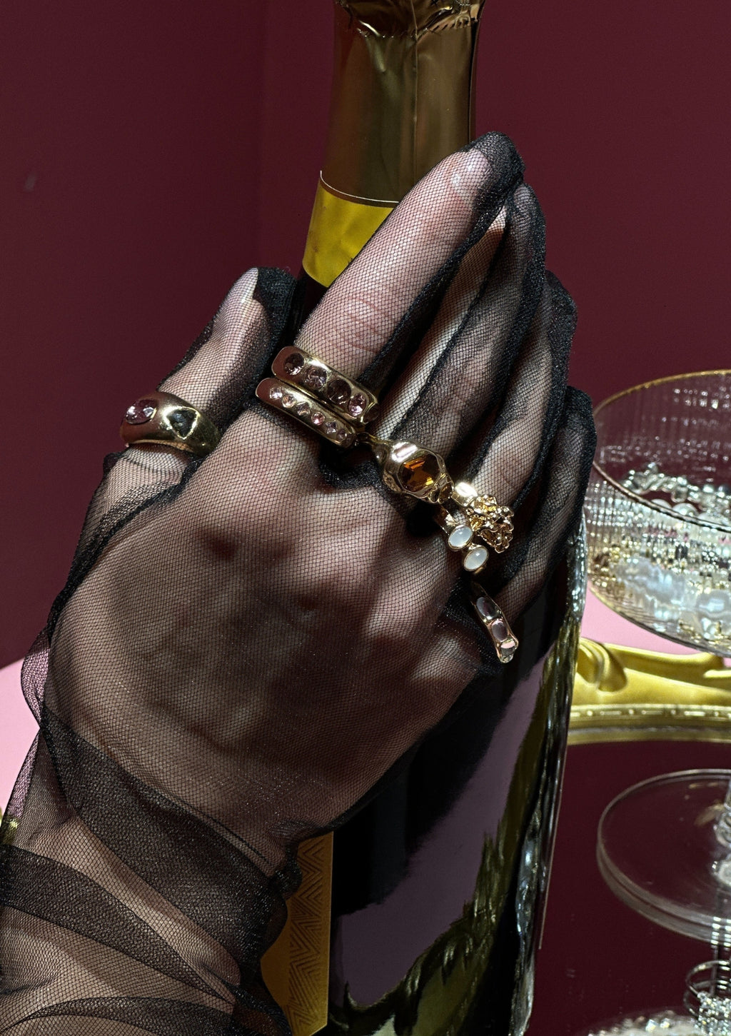 Hand wearing a sheer glove with multiple rings holding a bottle, with a red background.