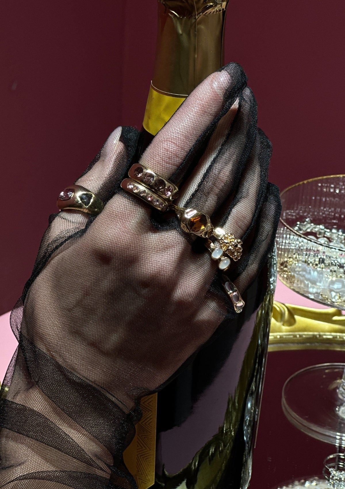 Hand wearing a sheer glove with multiple rings holding a bottle, with a red background.