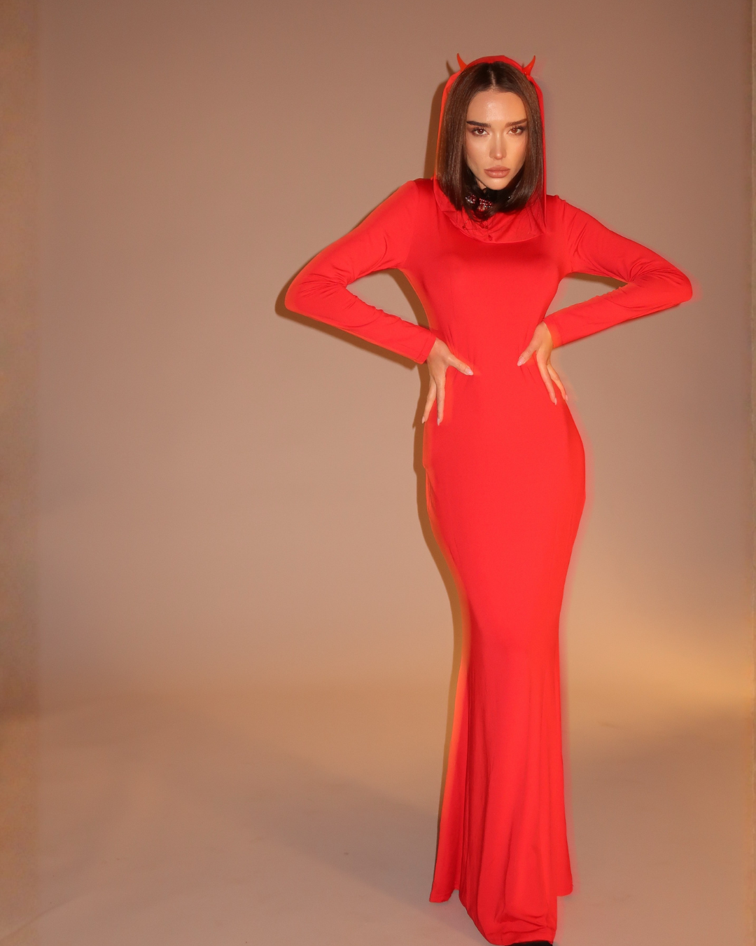 Person wearing a red dress with a hood and small horns on a beige background