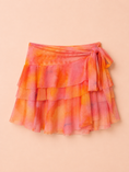 Заредете изображението в програмата за преглед на галерия, A tiered ruffle wrap skirt in a vibrant orange and pink tie-dye pattern, featuring a side tie closure, laid flat on a solid peach background.
