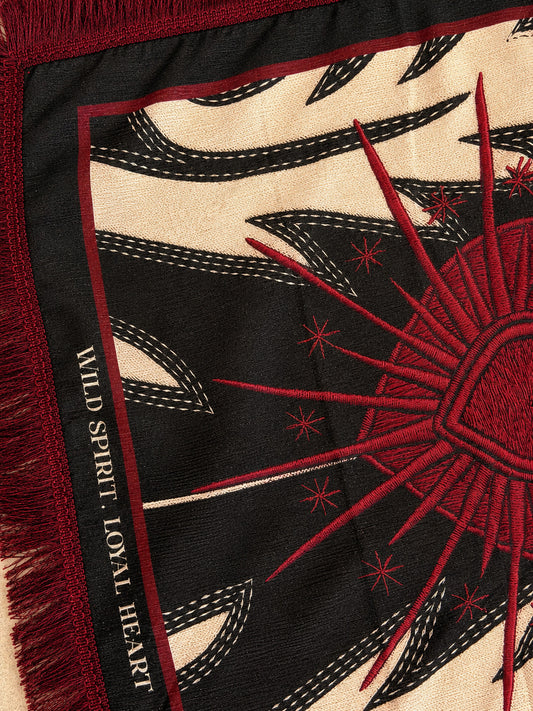 Close-up of a woven scarf with a bold sunburst design in red on a black and cream background, plus fringe and the words 'WILD SPIRIT. LOYAL HEART' along the edge.