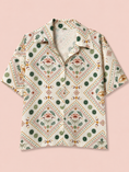 Заредете изображението в програмата за преглед на галерия, A short-sleeve button-up shirt with a bohemian-style geometric and floral pattern in green, orange, and brown on a cream background, displayed flat against a solid pale pink background.
