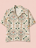 A short-sleeve button-up shirt with a bohemian-style geometric and floral pattern in green, orange, and brown on a cream background, displayed flat against a solid pale pink background.