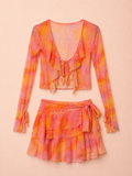A two-piece women's outfit set featuring a long-sleeve ruffled crop top and a matching tiered ruffled mini skirt, both in a vibrant orange and pink tie-dye pattern, displayed against a solid pale peach background.