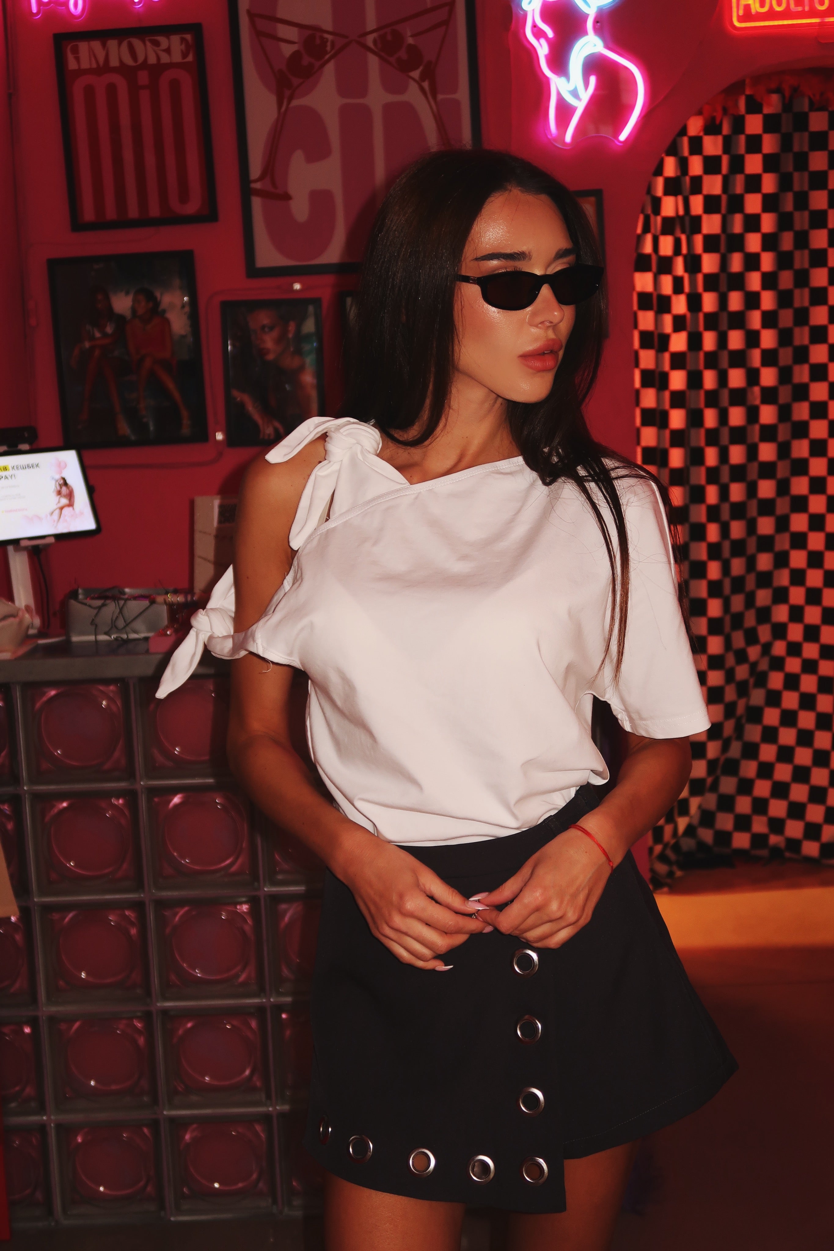 Woman in a white bow t-shirt and grey skirt standing in a room with neon signs and checkered patterns.