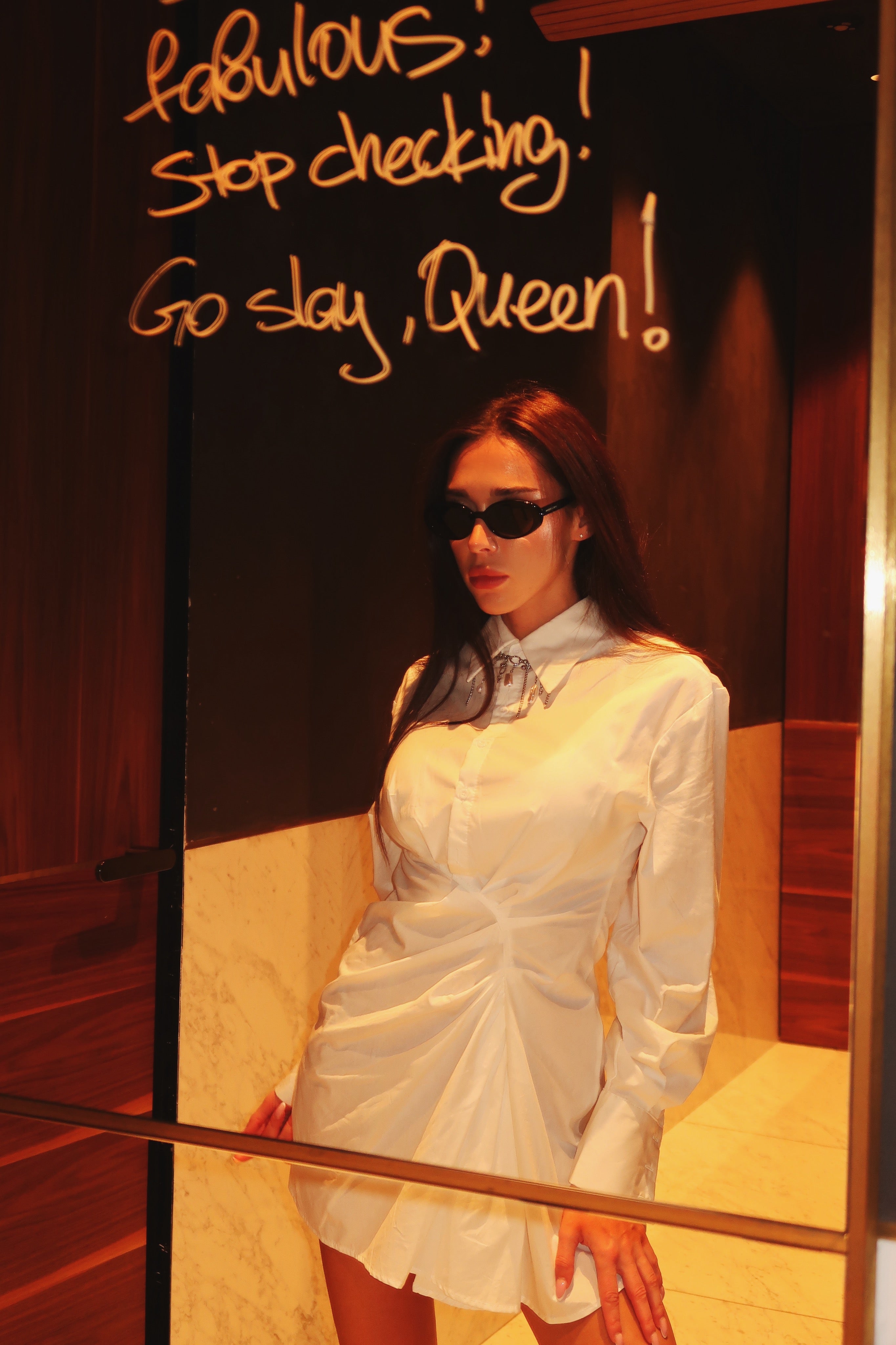 Woman in a white dress standing in front of a mirror with text on the wall.
