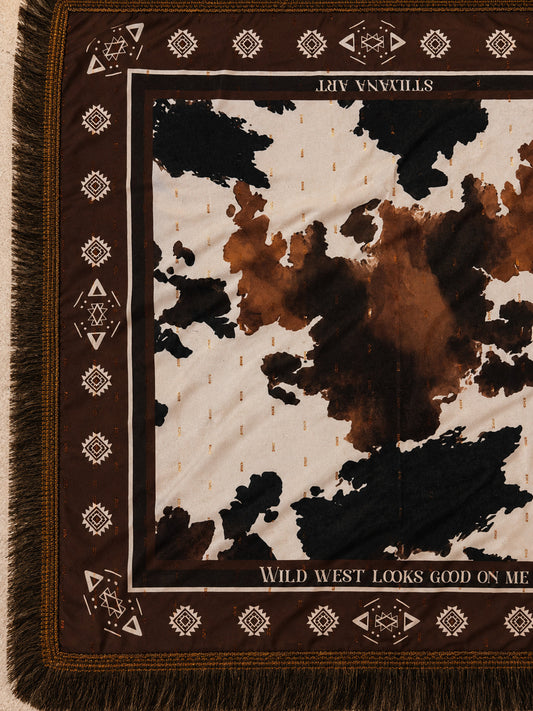A close-up of a woven scarf with fringe, featuring a brown and cream cow-print center surrounded by a dark brown border with geometric motifs and text reading 'STILL LIFE ART' and 'WILD WEST LOOKS GOOD ON ME'.